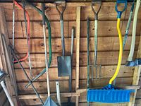Wide view showing two band saws, several shovels, a blue snow shovel with yellow handle, and garden tools hung on a wooden wall.