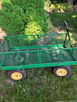 Side view of the green metal mesh garden wagon on grass showing mesh bed, tubular frame, side panels, wheels and pull handle partially visible