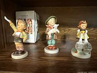 Three Hummel porcelain figurines displayed on a wooden shelf with original box in background.