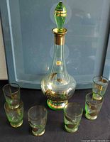 Full view of the green-tinted glass decanter with gold rose accents and six matching shot glasses arranged around it.