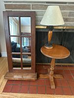 Wooden mirror with window pane frame and attached shelf, wooden round accent table, wooden table lamp with beige shade arranged in front of fireplace.