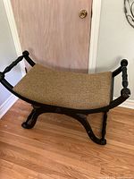 Full view of black and gold Regency style curved X base bench with beige upholstered seat on hardwood floor.