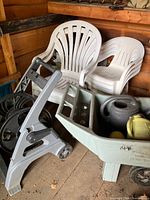 Six white plastic stacking chairs, plastic wheelbarrow with two watering cans inside, and a portable hose reel in a wooden shed.