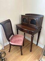 Full view of Queen Anne style wooden secretary desk with hutch and accompanying wooden chair with caned back and pink cushioned seat.