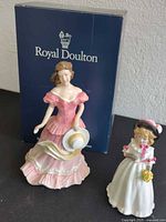 Two Royal Doulton figurines with blue branded box background. Woman in pink dress holding hat and a girl in white dress with flowers.