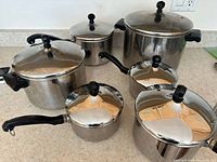 Set of 6 shiny stainless steel pots with lids showing varying sizes and handles, arranged on a light-colored countertop.