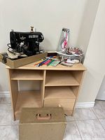 Vintage White sewing machine on beige base with attached power cord and foot pedal, placed on wooden cabinet with multiple open shelves.