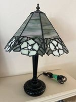 Full view of the stained glass lamp showing the greenish-grey and white floral-patterned glass shade and black base with cord coiled beside it.
