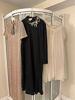 Three vintage garments hanging on a white clothing rack: beige snakeskin pattern dress, black dress with floral collar embellishment, and cream sheer dressing gown with crochet lace.