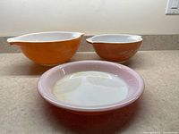 Wide view of two orange Pyrex mixing bowls with white interiors and one Pyrex pie plate with white interior and pink exterior on countertop.