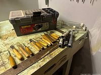 Wide view of woodworking tools and toolbox on a workbench, showing multiple carving chisels and gouges with wooden handles arranged in front of the toolbox.