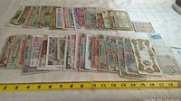 Two rows of assorted global bank notes laid flat with a measuring tape beneath them (approximately 12 to 36 inches visible).