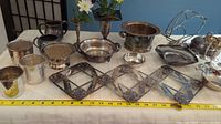Full view of silver ware collection with various bowls, urns, cups, trays, and expandable trivet rack with ruler for scale.