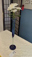 Full view of tall hand blown glass vase with white flowers inside, placed on light fabric surface with a decorative screen in background