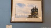 Framed watercolor painting showing a rocky shoreline and pine trees with calm water and a cloudy sky in the background.