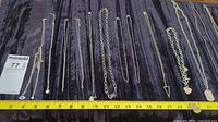 Multiple gold-tone and silver-tone chains laid out on fabric with measuring tape for length reference, showing various pendants including hearts, geometric shapes, and dog tag style pendants.