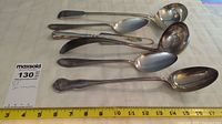 Seven assorted silver-tone serving spoons and ladles laid out side by side on a white cloth with a measuring tape below them showing lengths from 7 to 17 inches.