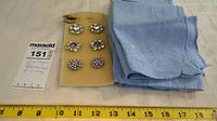 Vintage buttons in various rhinestone designs on yellow foam with linens stacked beside and a measuring tape below showing about 7 inches width