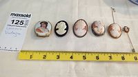 Six vintage cameos laid on fabric with a measuring tape showing scale around 3 to 8 cm. Includes painted porcelain brooch, four carved shell cameos, and one cameo stick pin.
