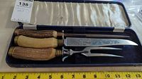 Photo showing the full vintage carving set in its blue case, including knife, fork, and sharpening steel with antler handles and etched metal blades.