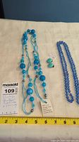 Two blue bead necklaces and turquoise blue earrings on a light background with yellow measuring tape showing length of necklaces