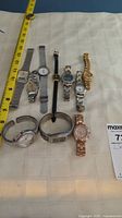 Lot of nine assorted wristwatches displayed on a fabric surface with measuring tape to side, showing variety of styles and band types.