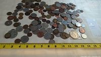 Many world coins spread out on a white surface with a measuring tape showing approximate length of 14 inches to indicate quantity and size.
