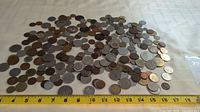 Top down photo showing a large pile of assorted world coins spread out with a ruler below showing scale.