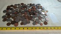Overhead wide shot of the large pile of assorted world coins arranged loosely on a cloth surface with a measuring tape showing approximately 14 inches wide spread.