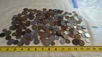 Wide view of assorted world coins spread on a white cloth with a yellow measuring tape showing approximately 15 inches length.