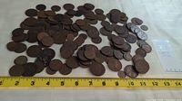 Wide view of scattered various world coins with a measuring tape below showing size reference, coins are mostly brown and aged.