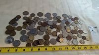 Wide view of various world coins of different sizes, colors, and denominations spread out on a table with rulers for scale.