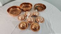 Overall view of 10 copper serving dishes: 3 hammered copper serving dishes and 7 small copper serving dishes with lids arranged on a white tablecloth.