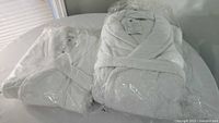Two white bath robes wrapped in clear plastic on a white surface near a window, one labeled large Velour Supima Cotton and other Terry Cotton