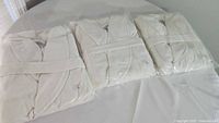 Three cream colored, large size robes, packaged in plastic, arranged on a white table surface. Each robe has a visible belt and logo tag confirming branded status.