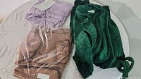Three garments on display: lilac dressing gown folded with tag, brown nightgown in plastic packaging, green robe laid open.
