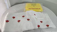 Four towels displayed on a table: large yellow promotional towel folded in packaging, large white bath towel with red floral embroidery unfolded on table, white hand towel and washcloth with matching floral embroidery folded beside bath towel.