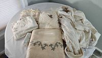 Top-down view of beige cream bath linen lot including terry robes, embroidered bath towel, pestemal beach towels, and wash cloths on round table.