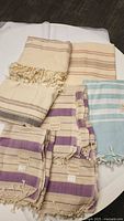 Photos showing seven Turkish cotton pestemal towels in assorted striped colors including beige, purple, and blue tones. Towels have fringe tassels.