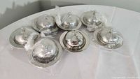 Photo showing all seven small hammered steel serving dishes with domed lids arranged on a white surface, with some lids wrapped in plastic.