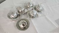 Seven small hammered steel finish serving dishes with dome-shaped lids on matching round plates, six of them covered in plastic wrapping, photo showing overall set.