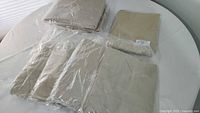 Nine beige/tan single flat fitted sheets, some wrapped in plastic, laid out on a white table.