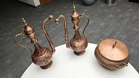 Three copper decorative pieces placed on white round table, two tall ewers and one round pot with lid