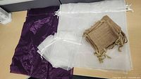Photo showing four large purple organza bags, eight large white organza bags, and twenty-three small burlap drawstring bags stacked.