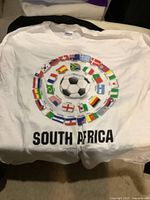 White Gildan T-shirt featuring a soccer ball graphic surrounded by flags of World Cup participating countries, with 'SOUTH AFRICA' text below.