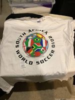 White Gildan T-shirt laid flat showing 2010 South Africa World Soccer event design with multi-national flags around a soccer ball.