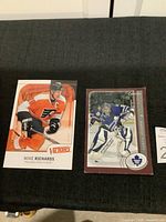 Photo showing two hockey trading cards, one of Mike Richards in Flyers uniform from Victory brand, the other Curtis Joseph in Leafs uniform from O-Pee-Chee brand, both laid on dark surface.