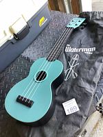 Front view of teal Kala The Waterman ukulele resting on a black textured surface next to a black gig bag with The Waterman logo and graphics.