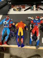 Photo showing two Spider-Man themed action figures on a table: one in blue suit with black spider emblem, one in red and blue armor with spider robot attachment.