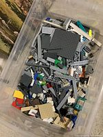 Clear plastic bin filled with a variety of LEGO pieces including grey structural parts, plates, and bricks